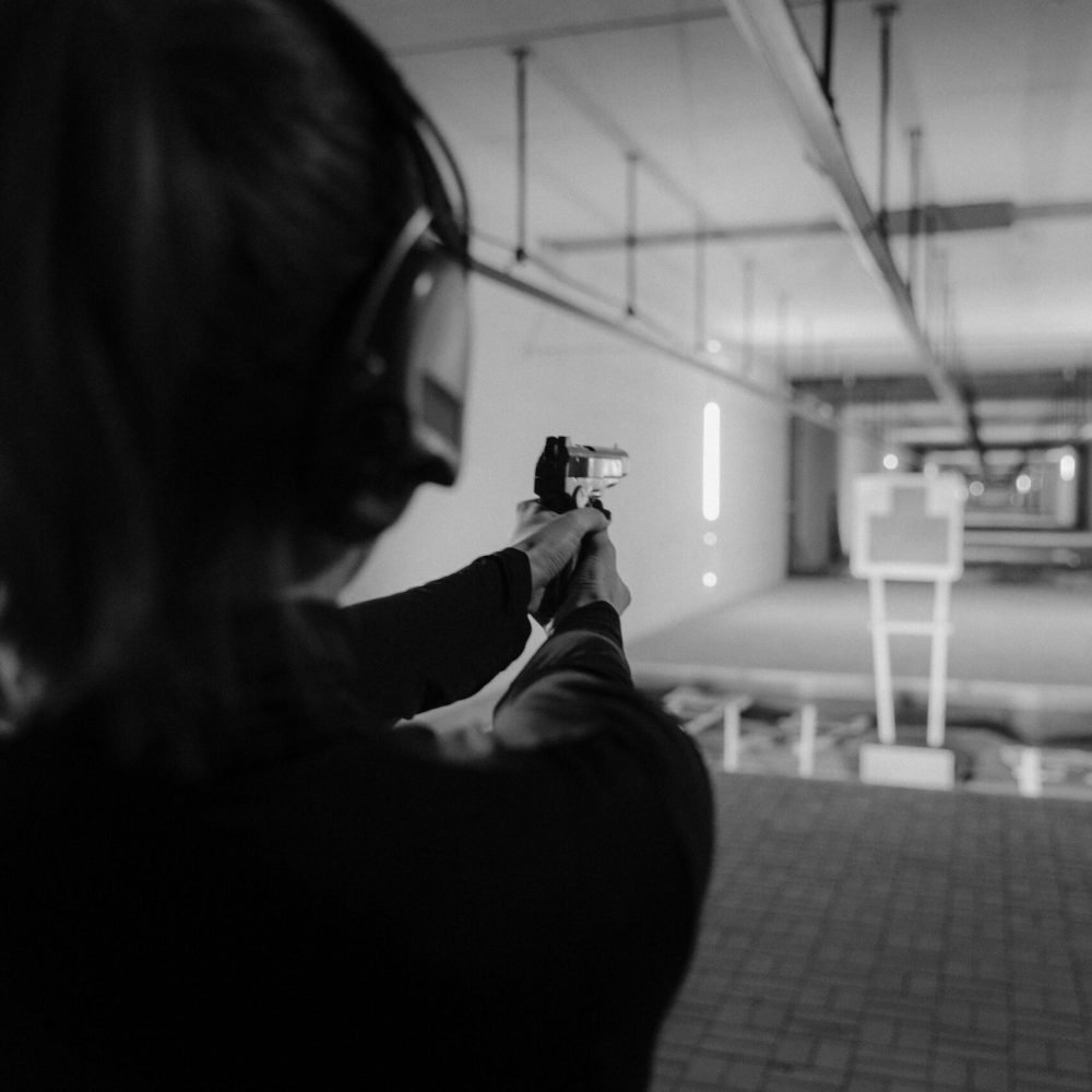 Black and white image of a woman shooting at an indoor range with headphones and pistol.